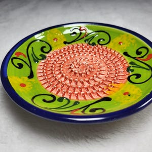 Puede incluir: Un plato rallador redondo de cer&aacute;mica que presenta una superficie central texturizada con crestas afiladas y elevadas en color rosa salm&oacute;n. El borde exterior est&aacute; pintado en verde lima brillante con patrones de remolinos negros, acentos amarillos y un borde azul oscuro.