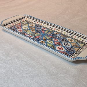 May include: A rectangular ceramic serving tray with a vibrant geometric pattern. The tray has a white base with a detailed design in blue, yellow, red, and green. The edges are trimmed with blue and black accents.