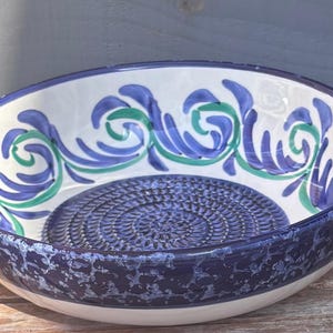 May include: A ceramic bowl with a white interior and a blue and green floral design around the rim. The bowl has a textured blue base and a dark blue speckled exterior.