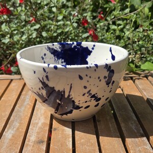 May include: A white ceramic bowl with dark blue paint splatters. The bowl is on a wooden surface, with a background of green foliage and red flowers. The bowl is suitable for serving food.