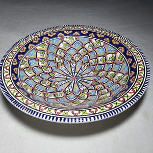 May include: A round ceramic bowl featuring a detailed, symmetrical mandala-style pattern in shades of blue, green, red, and yellow. The interior displays a repeating petal motif radiating from the centre, framed by a thick blue and white border.