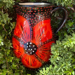 May include: A ceramic jug with a vibrant red poppy design. The jug features a black handle and rim, adorned with red dots. The poppy has a black centre with white dots and black lines radiating outwards. The jug is surrounded by green foliage.