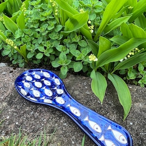 May include: Blue ceramic spoon rest with a white fish design. The spoon rest is sitting on a rock covered in green grass.