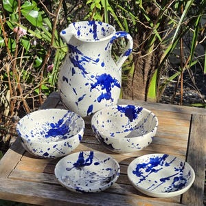 May include: A white ceramic jug and bowls set with a blue splatter design. The jug has a handle and a pouring spout. The bowls are of varying sizes, with a similar blue splatter pattern. The set is displayed on a wooden surface.