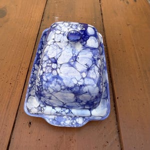 May include: A rectangular ceramic butter dish featuring a white base with a vibrant blue bubble-like pattern across the lid and the tray. The lid includes a small, rounded knob handle at the top, and the tray has slightly raised edges for containment.