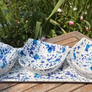May include: A set of three square ceramic bowls with rounded corners and a matching rectangular tray, all featuring a white base with a vibrant blue paint splatter pattern. The bowls are arranged in a row on the tray.