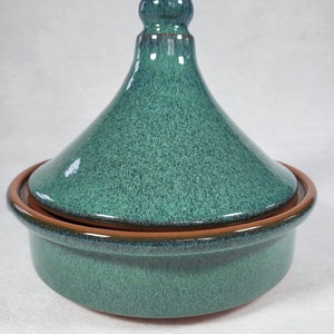 May include: A teal ceramic tagine pot with a conical lid and a rounded base. The lid has a small knob at the top. The rim of the base and lid are trimmed with a brown band. The pot has a speckled, glazed finish.
