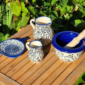 May include: A set of blue and white ceramic kitchenware, including a small pitcher, a larger pitcher, a small dish with a handle, and a mortar and pestle. The mortar and pestle have a blue rim and a white base with a blue floral pattern.