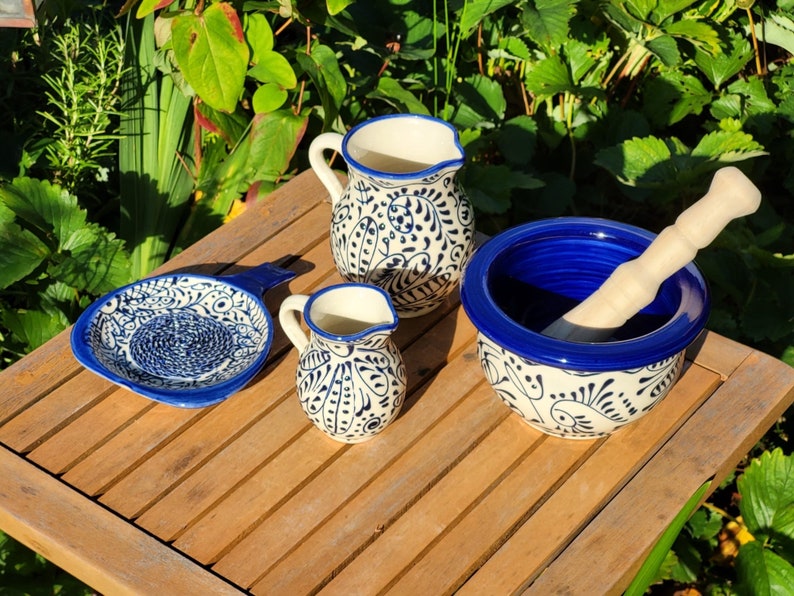 May include: A set of blue and white ceramic kitchenware, including a small pitcher, a larger pitcher, a small dish with a handle, and a mortar and pestle. The mortar and pestle have a blue rim and a white base with a blue floral pattern.