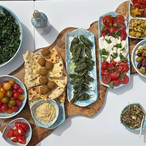 May include: A selection of dishes including a bowl of greens, cherry tomatoes, falafel with pita bread and hummus, and a platter of tomatoes and mozzarella. Other dishes include olives, peppers, and a grain salad, all arranged on a wooden board.