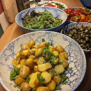 May include: A selection of dishes on a table, including a bowl of potatoes with mint, a salad, and tomatoes. The potatoes are in a blue and white patterned bowl. An olive oil bottle and other food items are also visible.