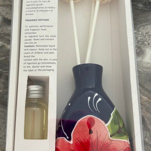 May include: A blue and red ceramic diffuser bottle with a floral design. The bottle is filled with a clear liquid and has two white rattan sticks. The bottle is in a white box with the text "Difusor de Fragancia" and "Fragance Diffuser" on it.