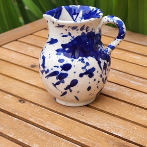 May include: A small white ceramic jug with a blue splatter paint design. The jug has a rounded body and a handle.