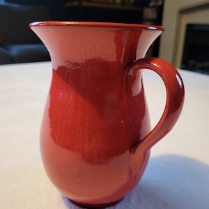 May include: A red ceramic jug with a wide mouth and a rounded body. The jug has a single handle.