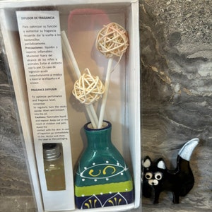 May include: A white box with a green and blue ceramic diffuser bottle, three brown wicker sticks, and a small black ceramic cat figurine. The box has text in Spanish that reads "Difusor de Fragancia" and "Precauciones: liquidos y vapores inflamables".