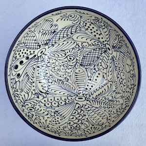 May include: A ceramic bowl with a cream-coloured interior and a dark blue rim. The bowl features an intricate hand-painted design in dark blue, showcasing floral and abstract patterns. The design is symmetrical and covers the entire interior surface.