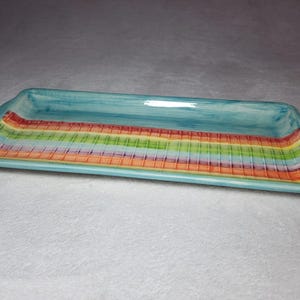 May include: A rectangular serving tray with a light blue glaze and a colourful striped design. The stripes are in shades of orange, yellow, green, and pink.