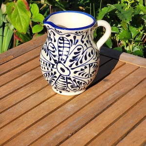 A white ceramic jug with a blue rim and intricate blue floral designs. The jug has a handle and is sitting on a wooden table.