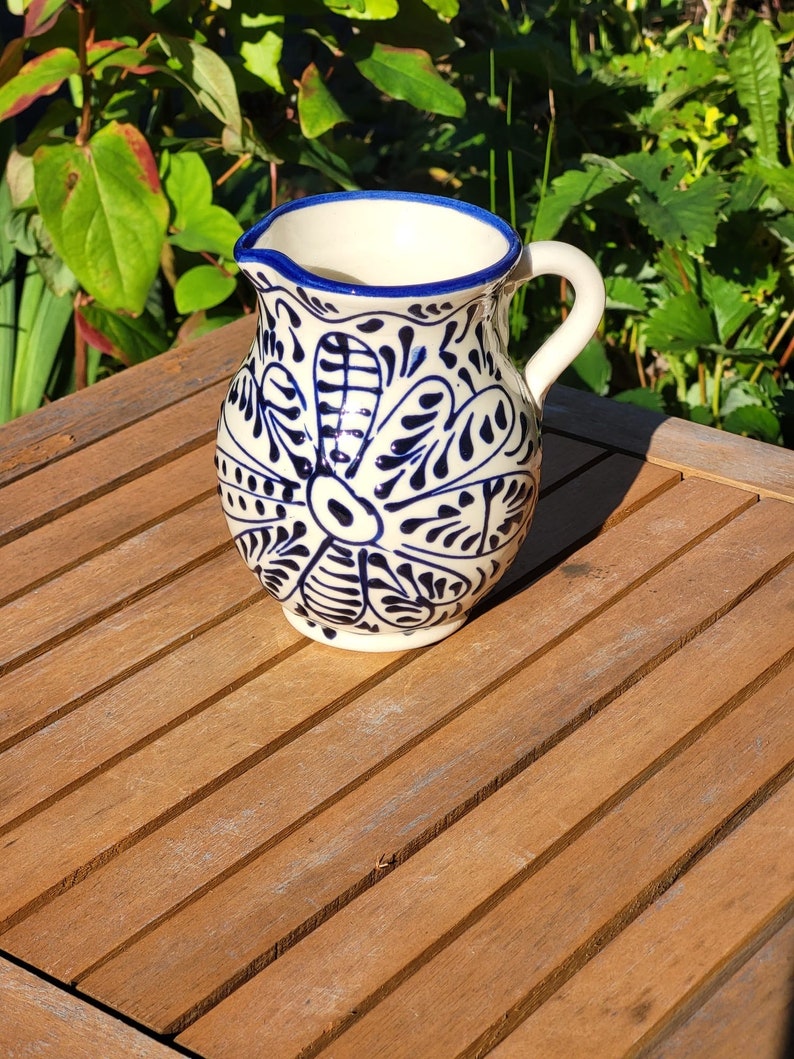 A white ceramic jug with a blue rim and intricate blue floral designs. The jug has a handle and is sitting on a wooden table.
