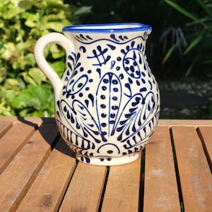 A white ceramic jug with a blue rim and intricate blue floral designs. The jug has a handle and is sitting on a wooden surface.