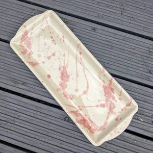 May include: Cream-coloured rectangular ceramic serving platter with pink splatter design. The platter has curved handles at each end. The pink paint is applied in a random pattern across the surface of the platter.