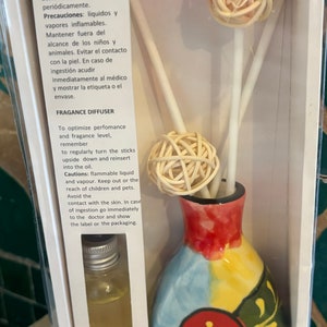 May include: A white box with a clear plastic window showing a colourful ceramic vase with a red flower design. The vase contains three wooden sticks with a ball of woven reeds on top. The box also contains a small bottle of clear liquid. The text on the box reads "Difusor de Fragancia" and "Fragance Diffuser".