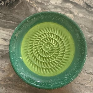 Puede incluir: Un plato rallador redondo de cer&aacute;mica con un centro de color verde lima vibrante y una textura en espiral elevada para rallar. El borde ancho es de un verde bosque m&aacute;s oscuro con motas blancas en el esmalte, creando un acabado moteado.