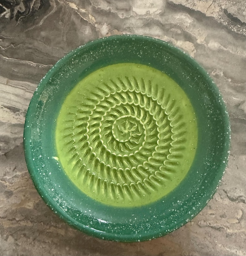 Puede incluir: Un plato rallador redondo de cer&aacute;mica con un centro de color verde lima vibrante y una textura en espiral elevada para rallar. El borde ancho es de un verde bosque m&aacute;s oscuro con motas blancas en el esmalte, creando un acabado moteado.