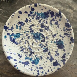 Puede incluir: Un plato rallador de cer&aacute;mica redondo con base blanca y un patr&oacute;n moteado de salpicaduras de pintura azul oscuro y azul claro. El centro del plato tiene una textura en espiral elevada dise&ntilde;ada para rallar alimentos como ajo o jengibre.