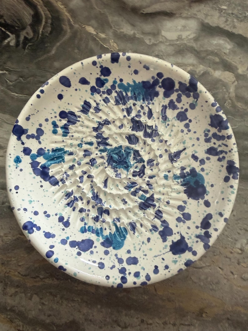 Puede incluir: Un plato rallador de cer&aacute;mica redondo con base blanca y un patr&oacute;n moteado de salpicaduras de pintura azul oscuro y azul claro. El centro del plato tiene una textura en espiral elevada dise&ntilde;ada para rallar alimentos como ajo o jengibre.