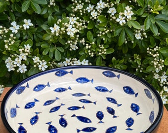May include: A white ceramic serving bowl with a blue rim and a pattern of blue fish. The bowl is sitting on a wooden surface.