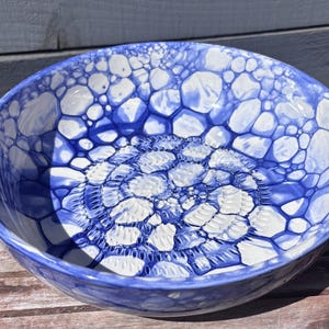May include: A round ceramic bowl featuring a vibrant blue and white bubble-like pattern across its interior surface. The centre of the bowl displays a textured, circular design with small, repeating indentations, surrounded by larger, organic shapes.
