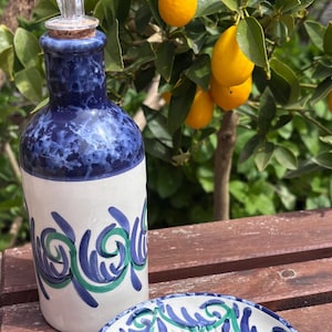 May include: A ceramic oil bottle and grater set with blue and green decorative patterns. The bottle has a clear spout and cork. The grater is white with a blue and green spiral design. Lemons grow on a tree in the background.