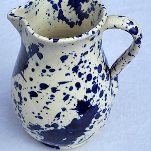 May include: A ceramic jug with a cream base and a striking blue splatter design. The jug features a curved handle and a spout for easy pouring. This decorative piece is ideal for serving beverages.