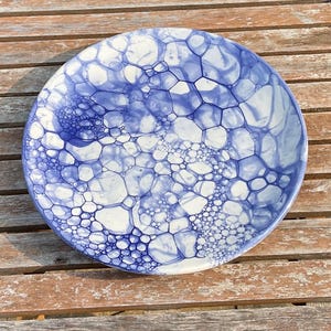 May include: A round ceramic plate featuring a white base colour with a blue bubble-like pattern across the surface. The design consists of interconnected circular shapes of varying sizes, creating a dense, organic network of blue lines and white spaces.