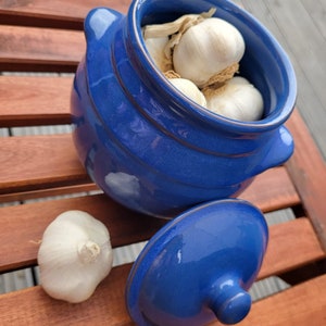 May include: A blue ceramic garlic keeper with a lid. The keeper is filled with garlic cloves. The keeper is sitting on a wooden surface.