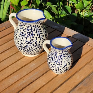 May include: Two ceramic pitchers with blue and white floral designs. The larger pitcher has a wider mouth and a handle. The smaller pitcher has a narrower mouth and a handle.