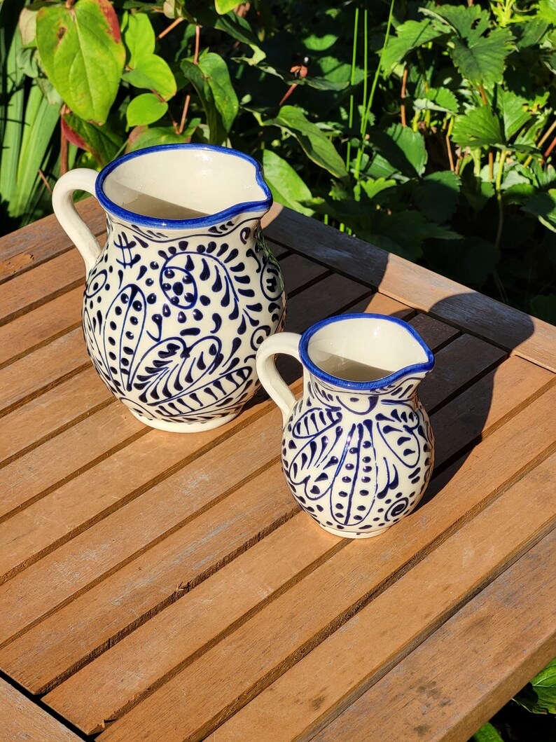 May include: Two ceramic pitchers with blue and white floral designs. The larger pitcher has a wider mouth and a handle. The smaller pitcher has a narrower mouth and a handle.