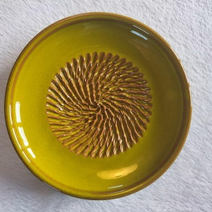 Puede incluir: Un plato rallador circular de cer&aacute;mica de color chartreuse con un borde liso y brillante y un patr&oacute;n en espiral texturizado y elevado en el centro, dise&ntilde;ado para rallar alimentos. El plato presenta un esmalte amarillo verdoso uniforme.