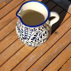 May include: A white ceramic pitcher with a blue and white floral design. The pitcher has a handle and is sitting on a wooden surface.
