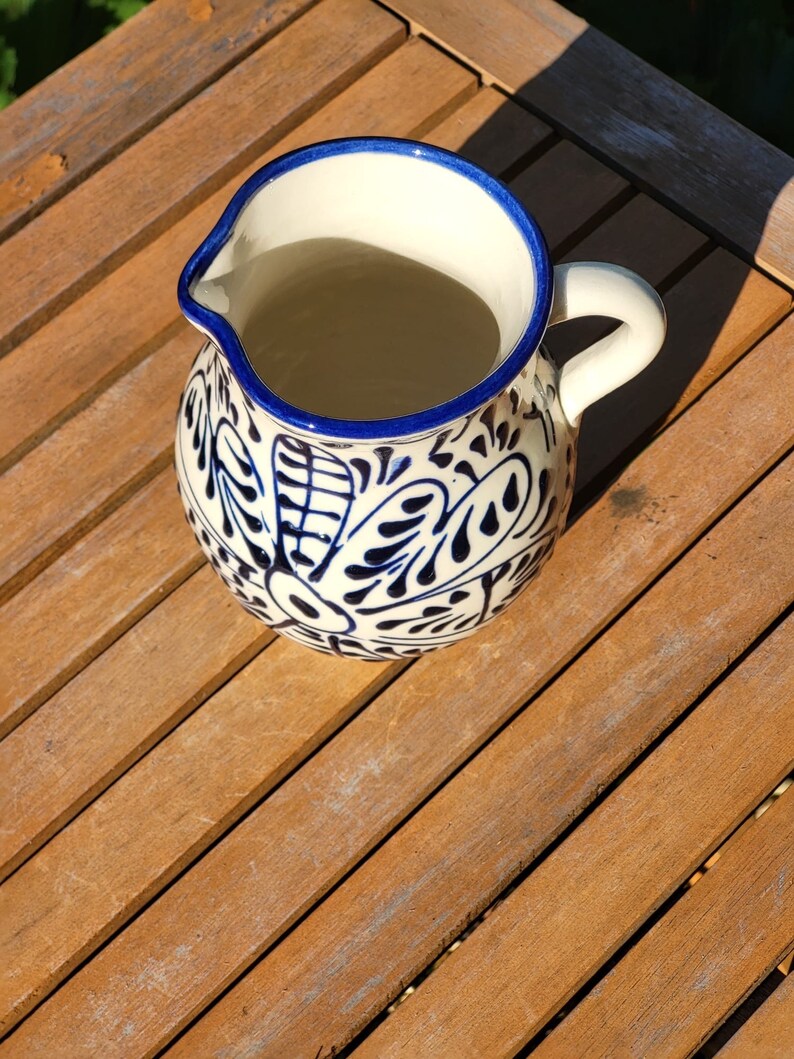 May include: A white ceramic pitcher with a blue and white floral design. The pitcher has a handle and is sitting on a wooden surface.