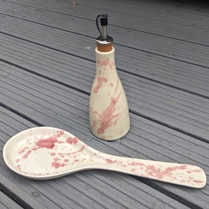 May include: A white ceramic oil bottle and spoon rest set with pink splatter design. The bottle has a stainless steel spout and cork stopper. The spoon rest is oval-shaped with a long handle. Both pieces are on a grey wooden surface.