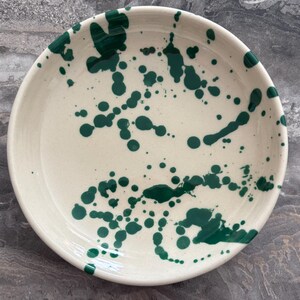May include: A round, off-white ceramic plate with a dark green splatter design. The plate has a slightly raised edge and a smooth surface. The green splatters vary in size and density, creating a unique, artistic look. Suitable for serving food.