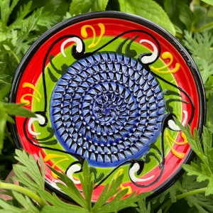 May include: A blue and white ceramic garlic grater with a red, green and yellow floral design. The grater is round and has a raised centre.