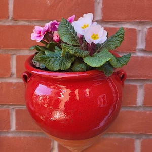 May include: A red ceramic wall planter with a white and pink flower arrangement. The planter has a rounded shape with a pointed bottom and two handles on the sides.