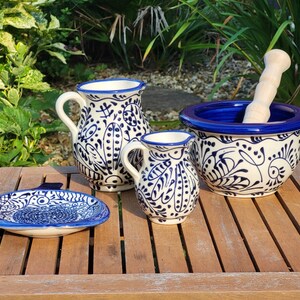 May include: A set of four blue and white Talavera pottery pieces, including a pitcher, a small cup, a mortar and pestle, and a small plate. The pieces are decorated with a traditional Mexican floral pattern.