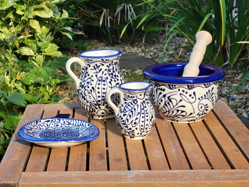 May include: A set of four blue and white Talavera pottery pieces, including a pitcher, a small cup, a mortar and pestle, and a small plate. The pieces are decorated with a traditional Mexican floral pattern.