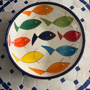 May include: A round ceramic bowl with a cream-coloured interior featuring a hand-painted pattern of nine stylised fish in various colours including orange, green, dark blue, red, yellow, and light blue, finished with a dark blue rim.