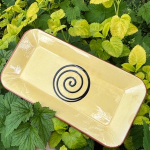 May include: A rectangular ceramic serving dish with a light yellow glaze and a black spiral design. The dish has a slightly raised rim.