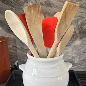 May include: A white ceramic utensil holder filled with wooden spoons and red silicone spatulas. The holder has two small handles and sits on a dark surface. The kitchen tools are arranged in a fan-like pattern.